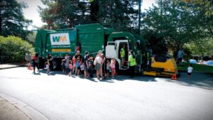 A large garbage truck parked with a line of adults and children waiting to climb inside